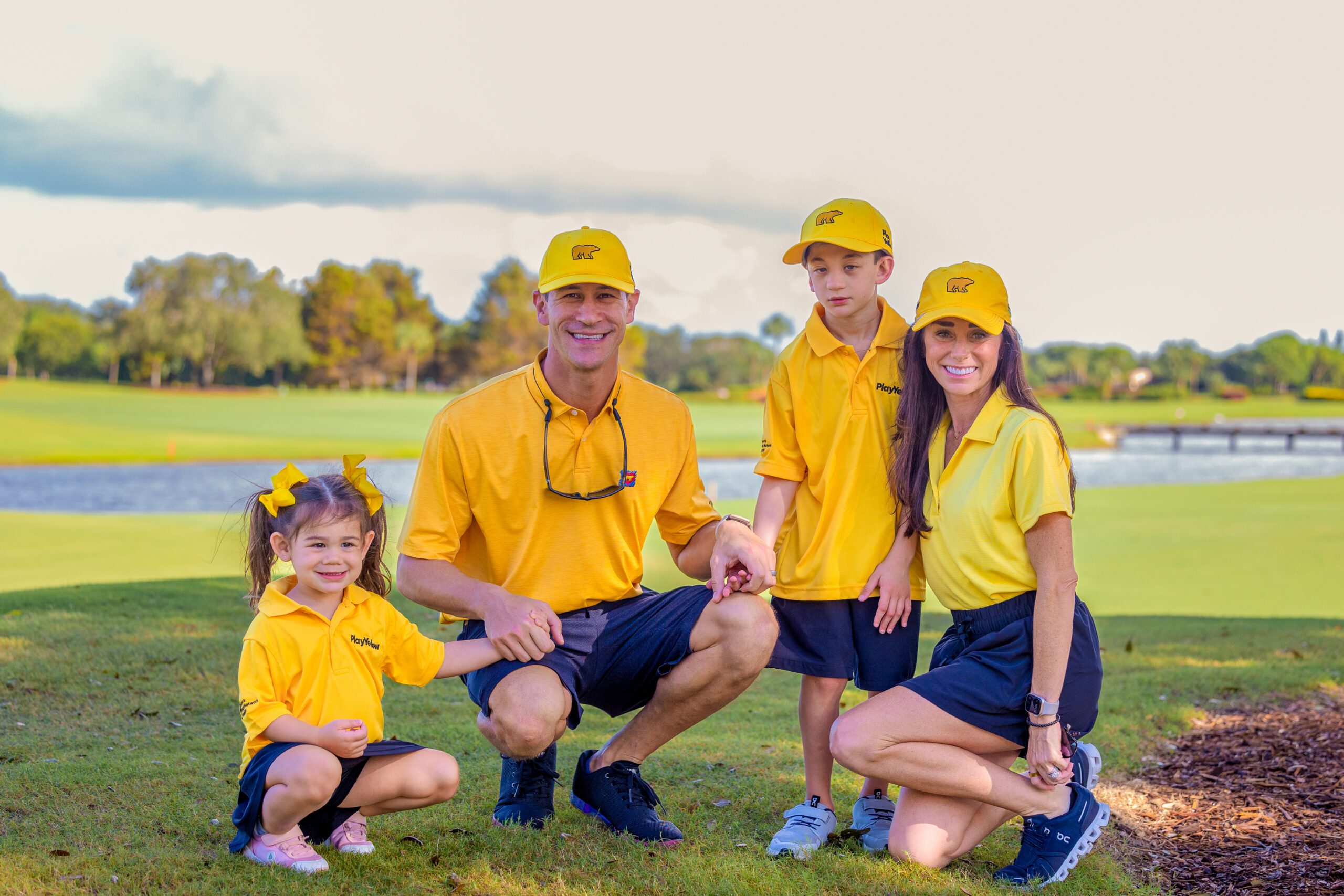 family of four, including two young children, posing on a golf course; everyone is wearing yellow Play Yellow polo shirts and yellow Golden Bear hats.