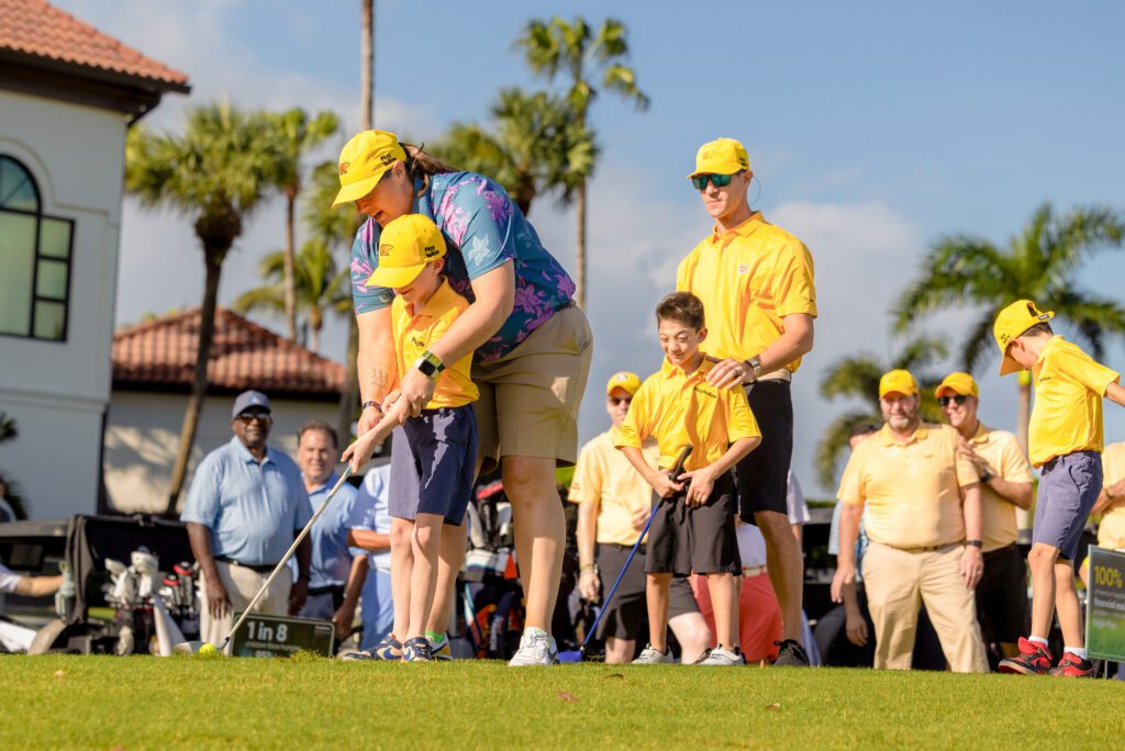A female golf instructor in a blue floral shirt and yellow Golden Bear hat assists a young boy in a yellow Play Yellow polo with his golf swing on a bright, sunny course. Several other participants and children, also unified by yellow Play Yellow apparel and hats, observe from the background near a clubhouse and palm trees.
