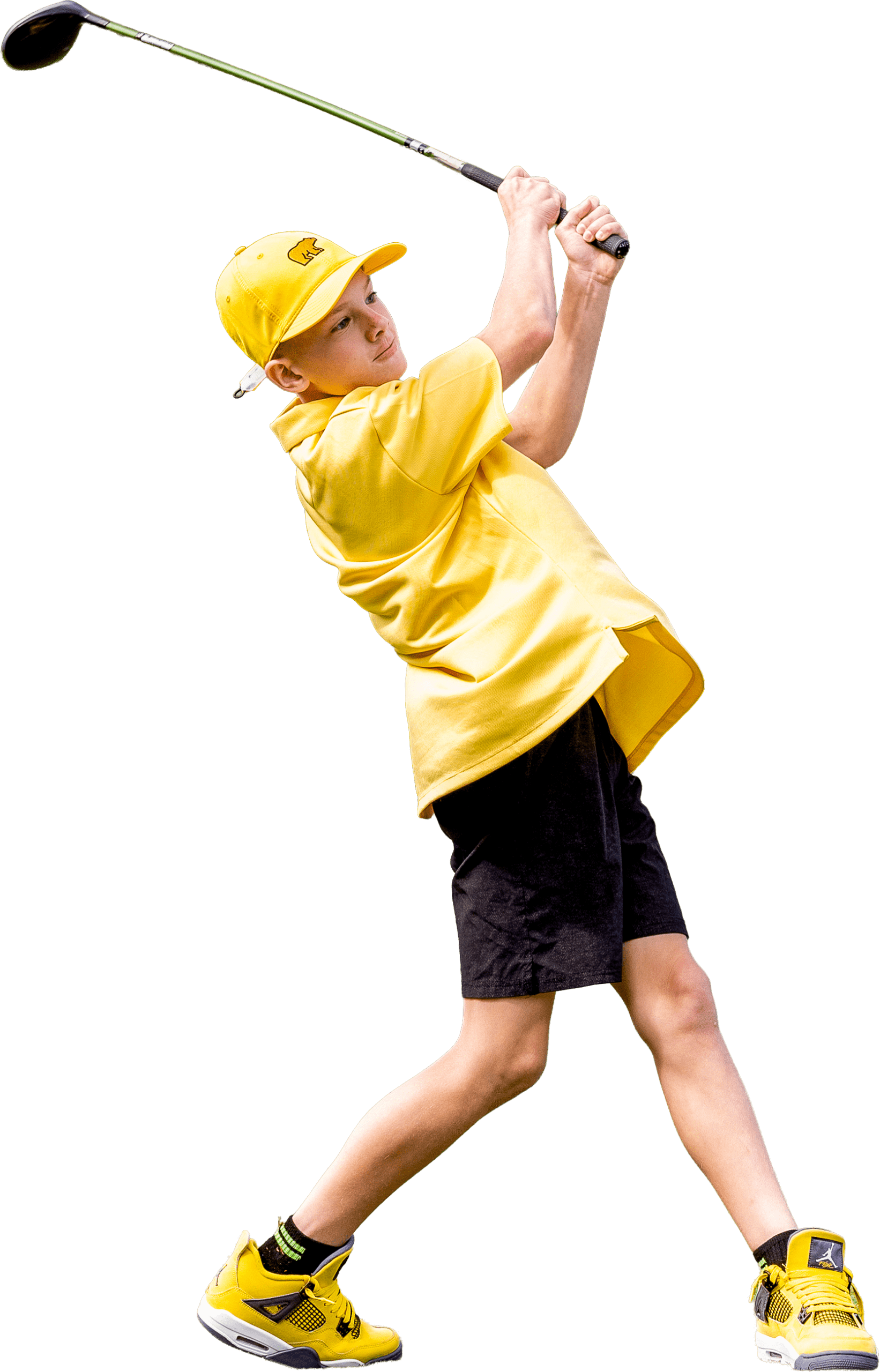 A young boy in a yellow polo shirt and a yellow Golden Bear hat completing a golf swing, set against a solid black background. He is wearing black shorts and vibrant yellow and black high-top sneakers.