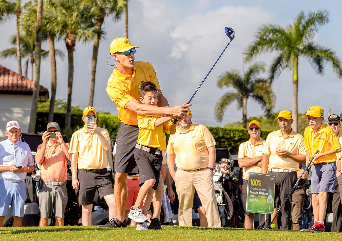Adult helping a young Play Yellow champion swing a golf club during an event.