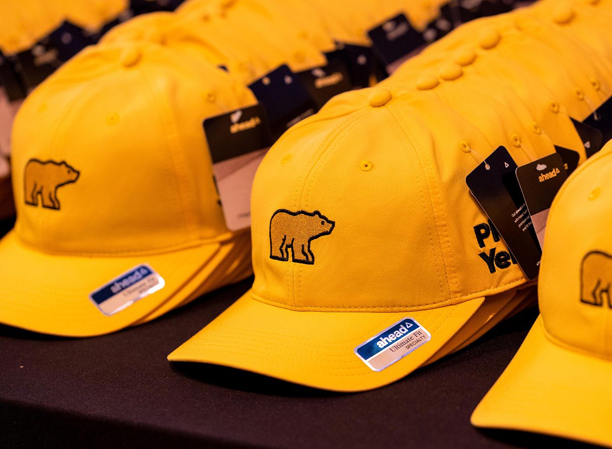 A row of bright yellow baseball caps by the brand AHEAD, featuring the black embroidered 'Golden Bear' logo of Jack Nicklaus on the front. The side of the hats are embroidered with the 'Play Yellow' campaign logo, and silver brand stickers are visible on the brims.