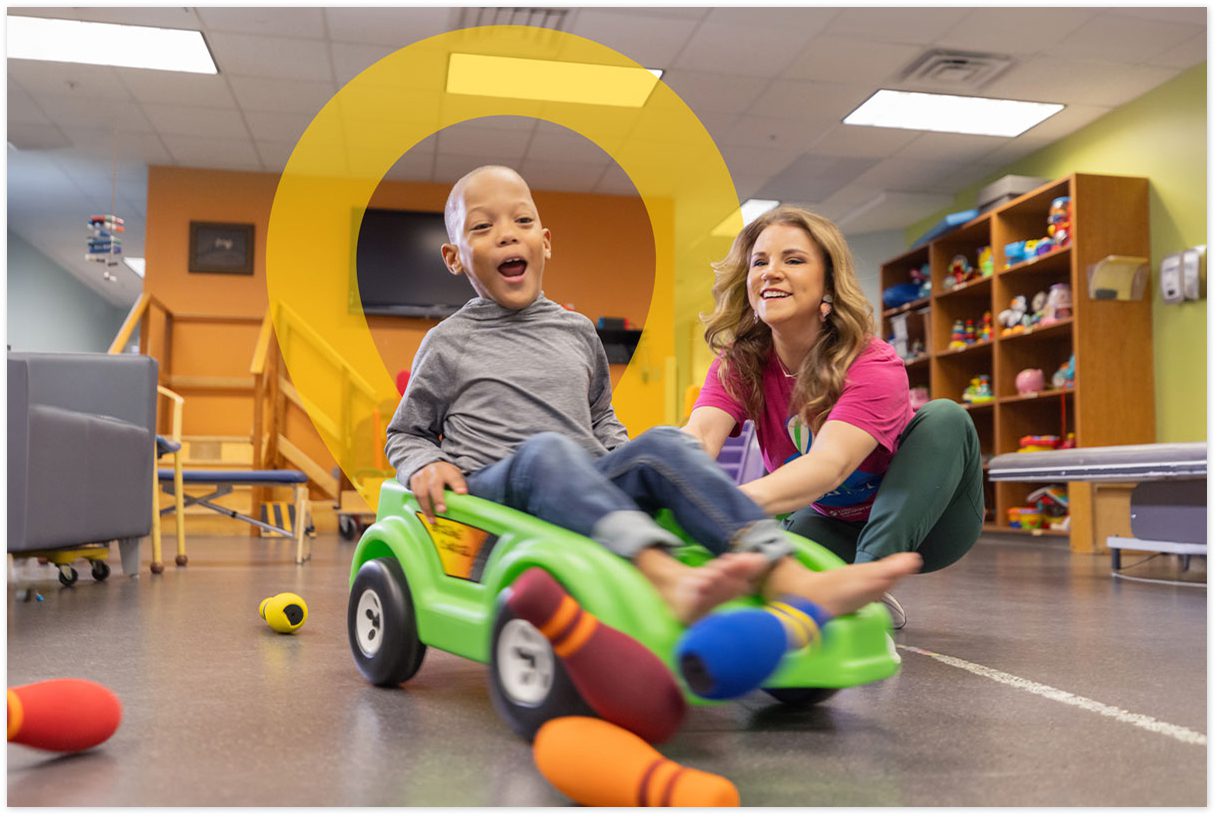 Child riding a toy car with support from a hospital staff member.