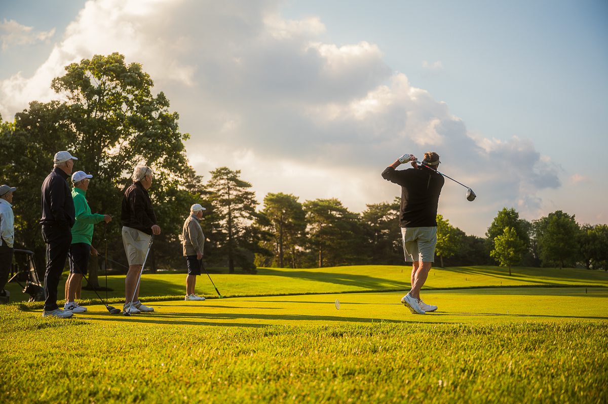 A group of senior golfers on a tee box watching a fellow player in a black long-sleeve shirt and grey shorts complete a drive during a golden-hour sunset.