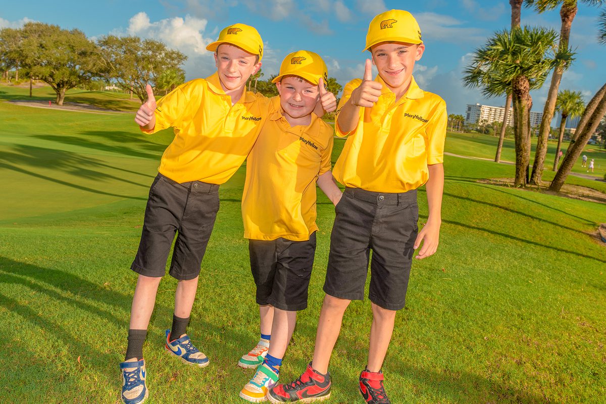 Three young boys wearing yellow Play Yellow polo shirts and yellow Golden Bear hats give thumbs-up gestures while posing together on a golf course.