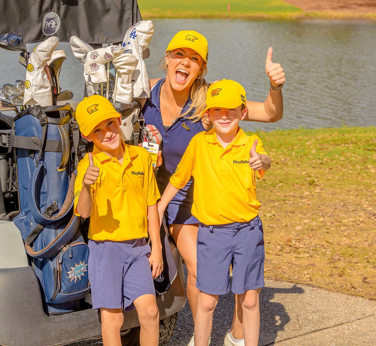 Two young golfers and an adult posing with thumbs up beside a golf bag at a Play Yellow event.