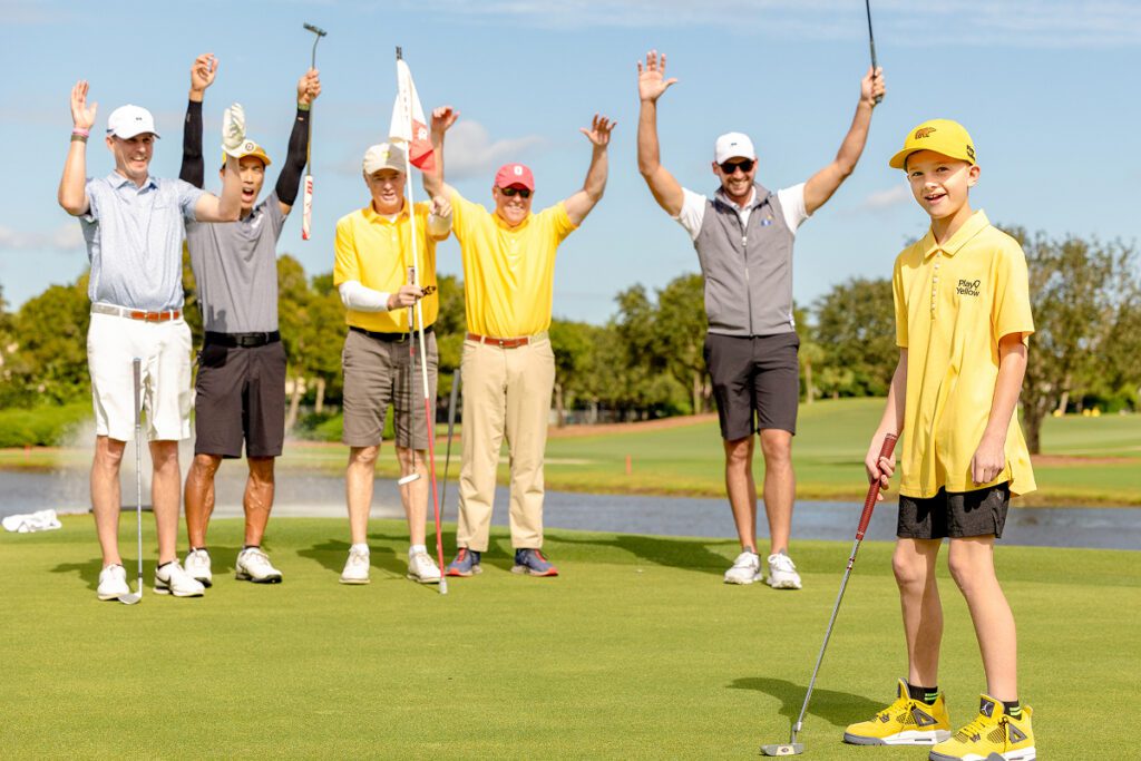 A young boy in a yellow Play Yellow polo and Golden Bear hat stands on a green with a putter while a group of adult golfers celebrates with their arms raised in the background.