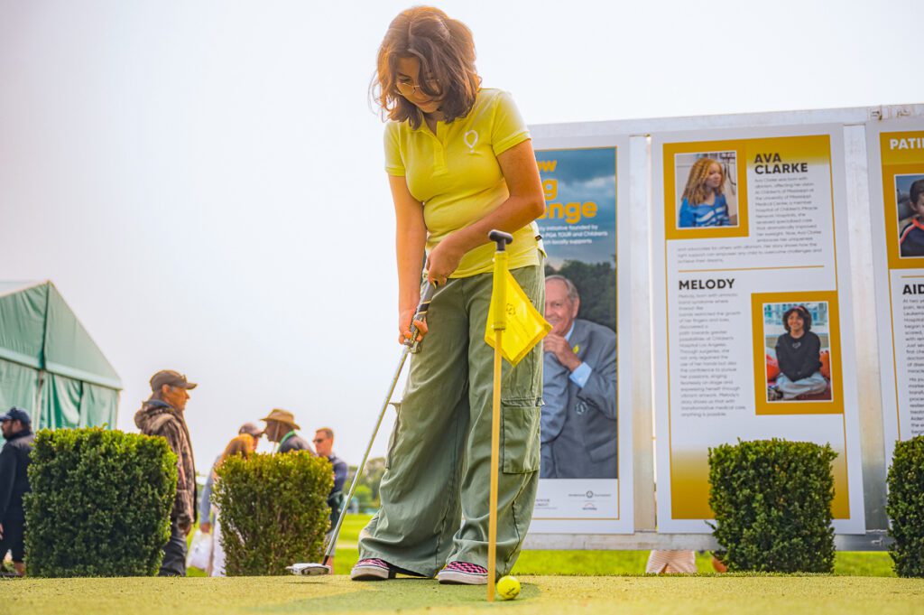 A young girl in a yellow Play Yellow polo shirt (Melody) and green cargo pants concentrate on a putt on a practice green, with informational Play Yellow posters featuring Ava Clarke and Melody in the background.