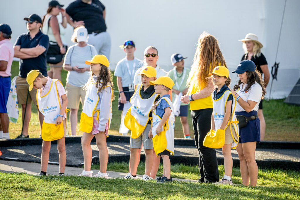 A line of children wearing yellow Play Yellow hats and white-and-yellow caddie bibs standing on the edge of a golf path during an event.