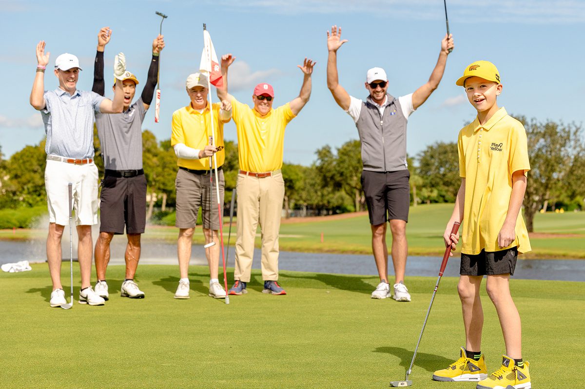A young boy in a yellow Play Yellow polo and Golden Bear hat putts on a green while a group of five men, including several wearing Play Yellow gear, cheer with their hands raised in the background.