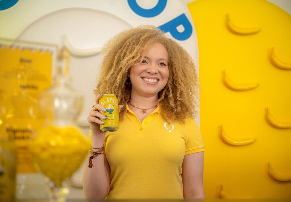 A smiling champion with curly hair wearing a yellow Play Yellow polo shirt, holding a can of yellow Olipop Banana Cream soda in front of a background decorated with yellow bananas.