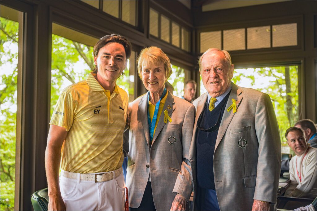 PGA Tour golfer Rickie Fowler in a yellow striped polo posing with Barbara and Jack Nicklaus, who are wearing grey blazers with Play Yellow ribbons pinned to their lapels.