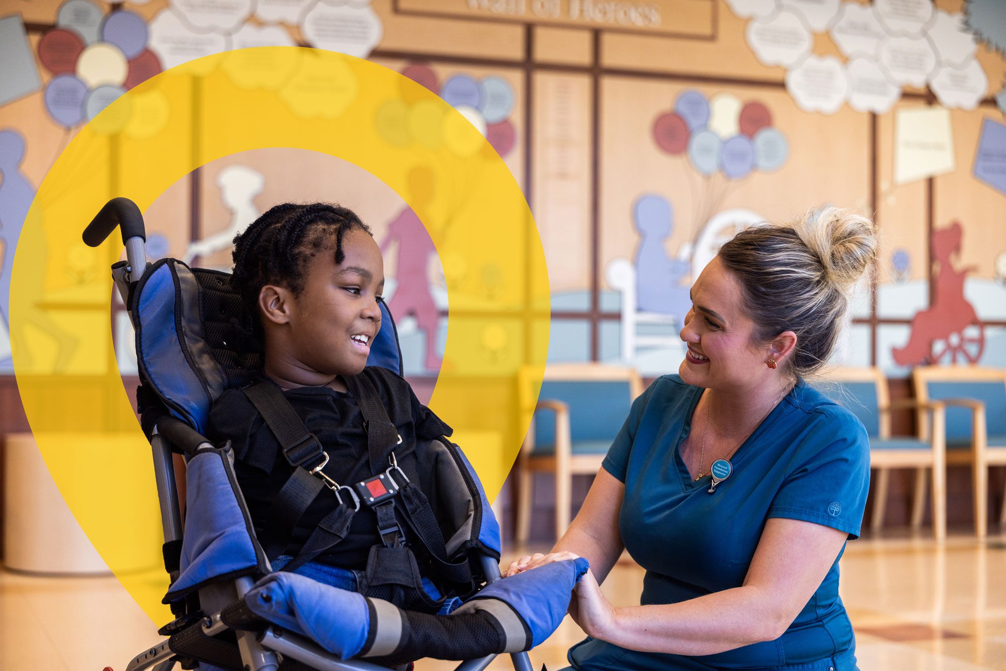 Child in a wheelchair smiling with a hospital staff member in a waiting area.