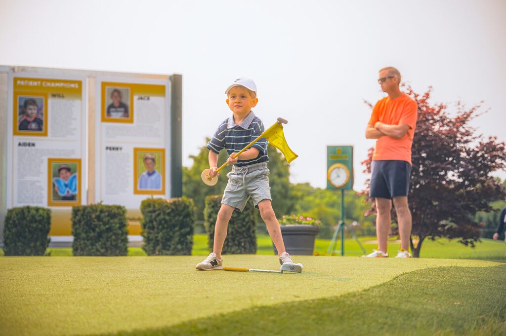 A young boy in a striped polo and white hat holds a yellow golf pin flag on a practice green, with a man in an orange shirt watching in the background.