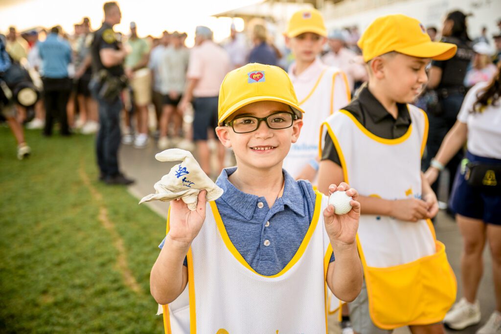 A smiling young boy wearing glasses and a yellow Play Yellow hat and bib holds up a white golf ball and a signed white golf glove for the camera.