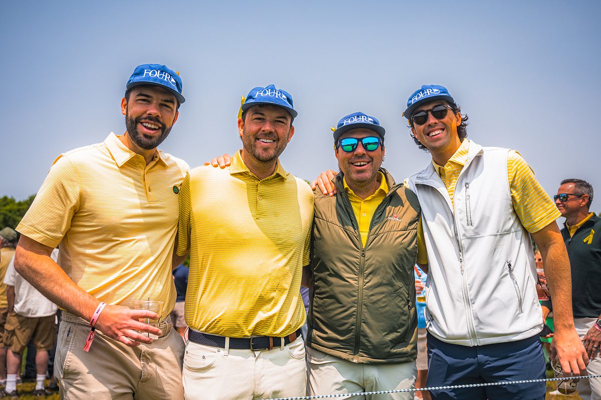 Four men pose together outdoors at a golf event, all wearing matching blue hats with white "FOUR" embroidery and yellow ribbons tucked into the side. They are dressed in variations of yellow apparel, including striped polo shirts, a green quilted vest, and a white performance vest, with several men holding plastic cups.