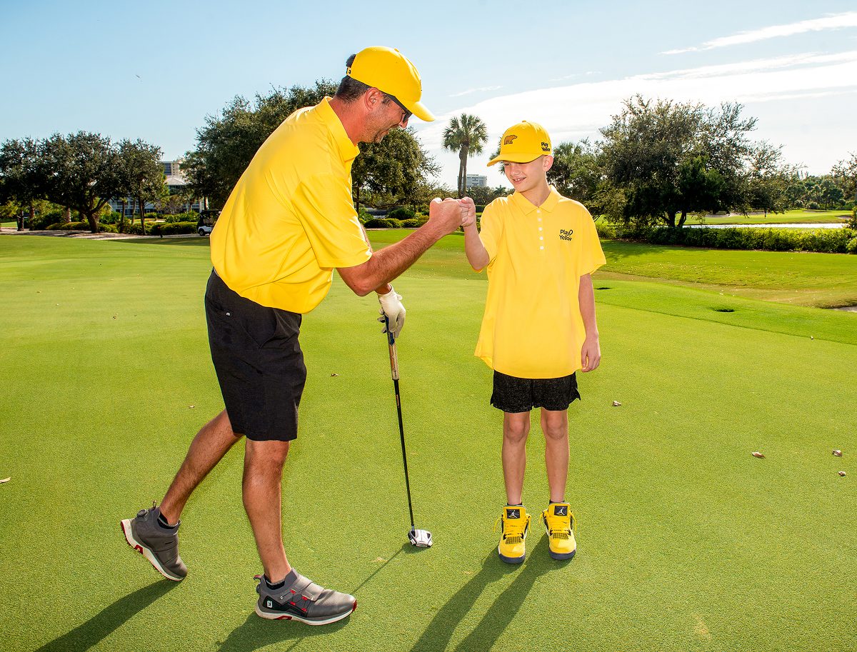 Play Yellow golfer fist-bumping a young participant on a golf course.